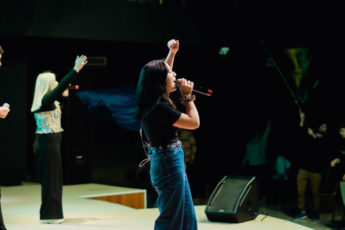 Energetic female musician singing live on stage with arm raised in front of an audience.