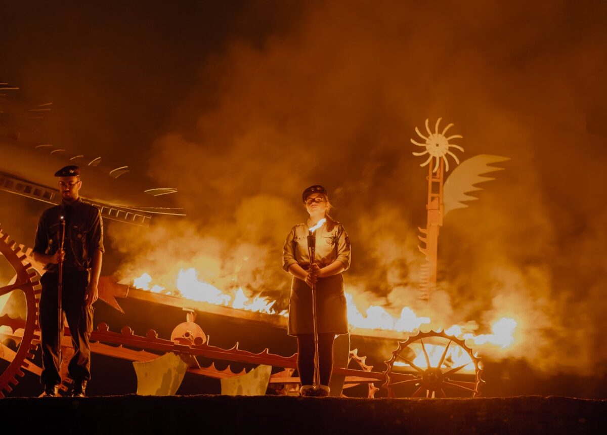 Two performers in vintage uniforms stand amidst dramatic flames and smoke in a theatrical setting.